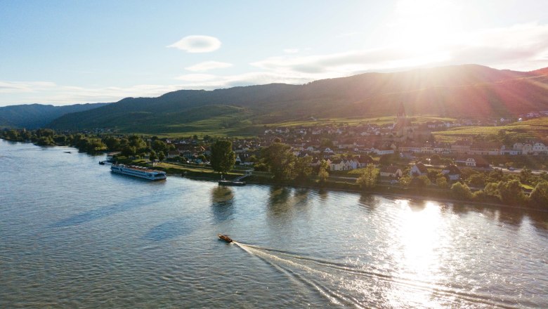 ahoi-wachau-Luftaufnahme, © NÖW_Daniel Gollner Luftaufnahme der Wachau mit Fluss, Booten und Dorf im Sonnenuntergang.