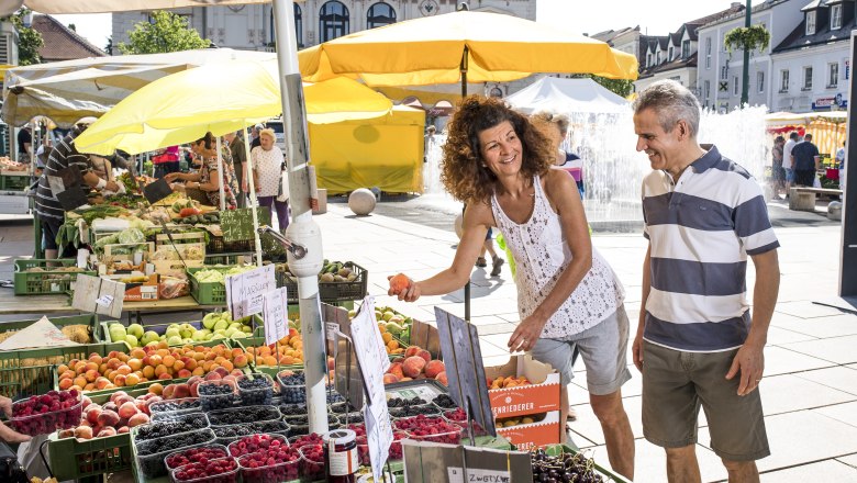 Tullner Naschmarkt, © Stadtgemeinde Tulln, Robert Herbst Menschen kaufen Obst auf einem Markt mit gelben Sonnenschirmen.
