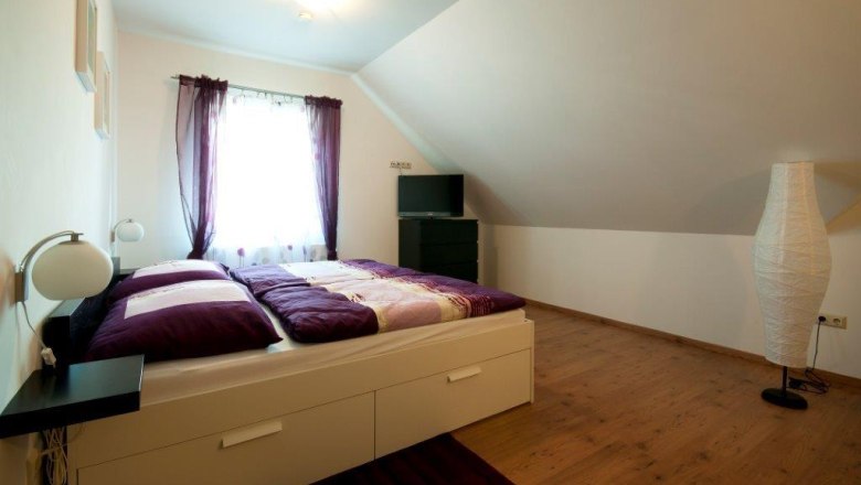 Gästezimmer, © Familie Rain Ein gemütliches Gästezimmer mit einem Doppelbett, lila Vorhängen und einem Fernseher auf einer Kommode unter einer Dachschräge.