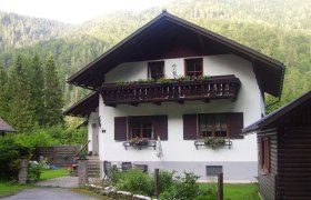 Ferienhaus Hager, © zVg Ein traditionelles Ferienhaus mit Holzbalkon und Blumenkästen, umgeben von grünen Bäumen und Bergen.