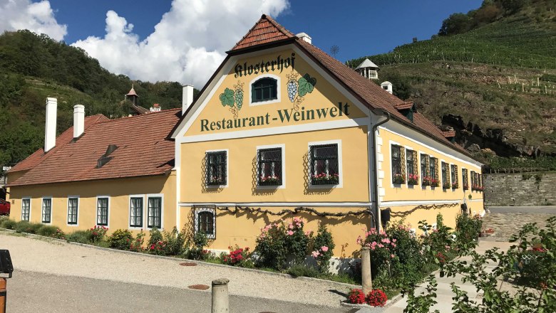 Aussenansicht vom Klosterhof, © Klosterhof Restaurant GmbH Außenansicht eines gelben Gebäudes mit der Aufschrift 'Klosterhof Restaurant-Weinwelt', umgeben von Blumen und Weinbergen.