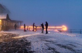 Bergadvent Rax, Advent, Raxalm-Bergstation, Winter, Rax-Seilbahn, Region Semmering-Rax, Wiener Alpen in Niederösterreich, © Wiener Alpen/Christian Kremsl Menschen stehen auf verschneiter Bergterrasse im Nebel, beleuchtet von warmen Lichtern bei Dämmerung.