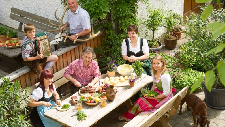 Zusammenkommen bei der Jause im Gastgarten, © Rita Newman Eine Familie in traditioneller Kleidung sitzt an einem Tisch im Garten und bereitet eine Mahlzeit vor.