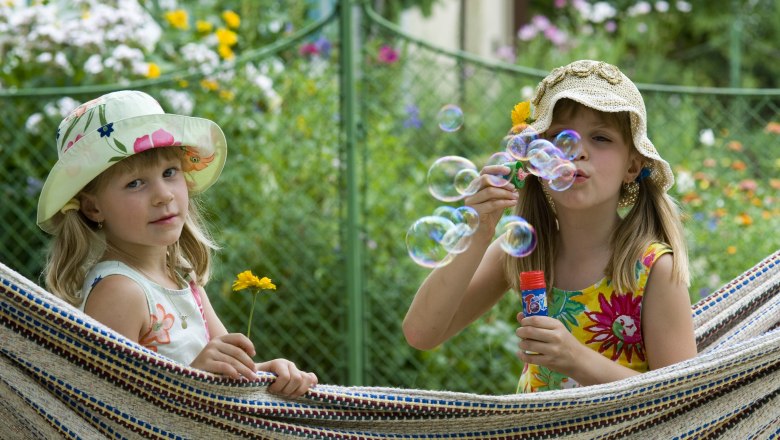 Kinder, © Weinviertel Tourismus GmbH / Himml Zwei Mädchen in Sommerkleidern und Hüten spielen in einer Hängematte im Garten. Eines hält eine Blume, das andere pustet Seifenblasen.