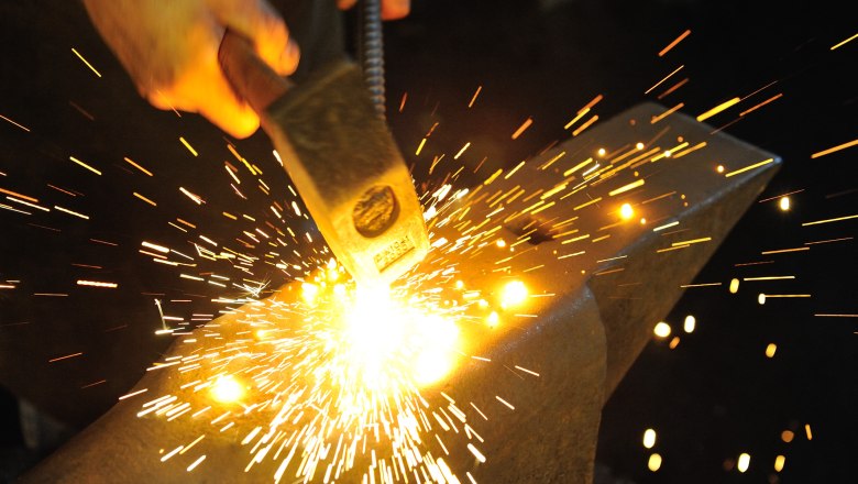 Arbeiten in der Hammerschmiede, © Gregor Semrad Ein Schmied schlägt mit einem Hammer auf glühendes Metall auf einem Amboss, Funken sprühen.