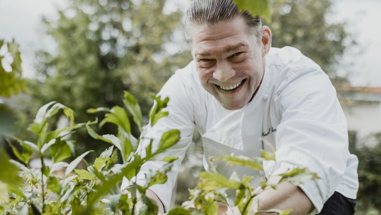 Küchenchef und Wirt Michael Böhm, © Niederösterreich Werbung/David Schreiber Lächelnder Koch in weißer Uniform im Garten, umgeben von Pflanzen.