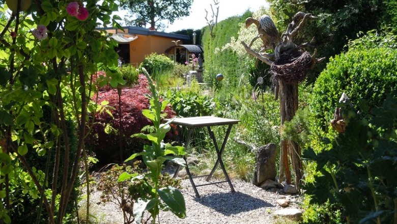 dscn5959_ausschn_c_die-gaerten-niederoesterreichs, © "Natur im Garten" Schaugärten Ein idyllischer Garten mit einem kleinen Tisch, umgeben von üppigem Grün und Blumen. Im Hintergrund ist ein gelbes Gebäude sichtbar.