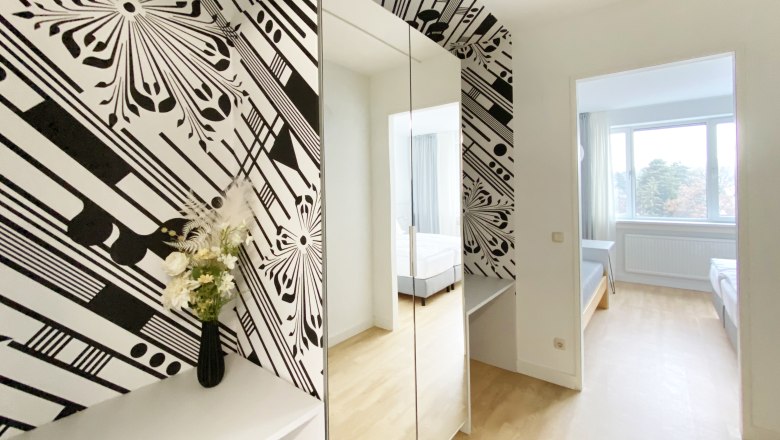 Gästezimmer, © Stadthotel Hollabrunn Modernes Gästezimmer mit schwarz-weißer Tapete, Spiegel, Blumen und Blick auf ein Bett durch eine Tür.