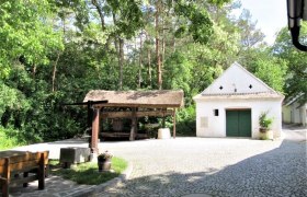 Weinviertler Kellergassenkulinarium, © D`Raschalaer Köllamauna Ein gepflasterter Hof mit einem kleinen weißen Gebäude und einem überdachten Holzgestell, umgeben von Bäumen.