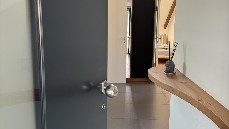 Eingangsbereich, © Juwel am Stadtrand Moderner Eingangsbereich mit offener Tür, Fliesenboden und Holzregal an der Wand.