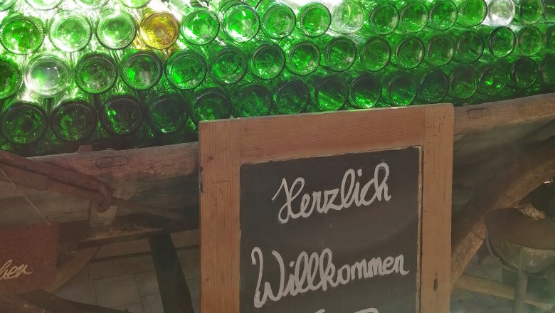 Weinbauernhof Wiesinger, © Andrea Wiesinger Ein Stapel grüner Glasflaschen auf einem Wagen mit einem Schild 'Herzlich Willkommen' davor, im Hintergrund ein Haus und Bäume.