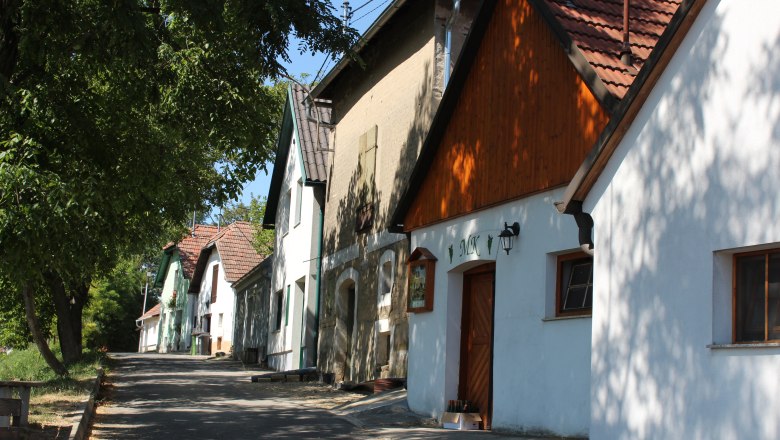 Weinviertler Kellergassenkulinarium, © Weinviertel Tourismus GmbH / Kroenigsberger Reihe von Weinkellern in einer ländlichen Straße mit Bäumen und blauem Himmel.