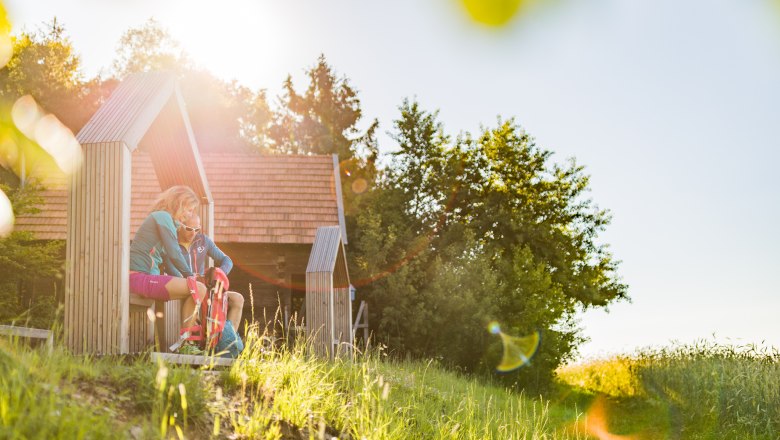 Rastplatz am Hutwisch mit Weitblick, © Wiener Alpen/Martin Fülöp Zwei Personen sitzen in einer hölzernen Struktur in einer sonnigen Landschaft.