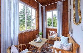 Top 1 - Wintergarten, © VP FeWo OG Ein gemütlicher Wintergarten mit Holzwänden, Korbmöbeln, blauen Vorhängen und einem Tisch mit Kerze und Blumen.