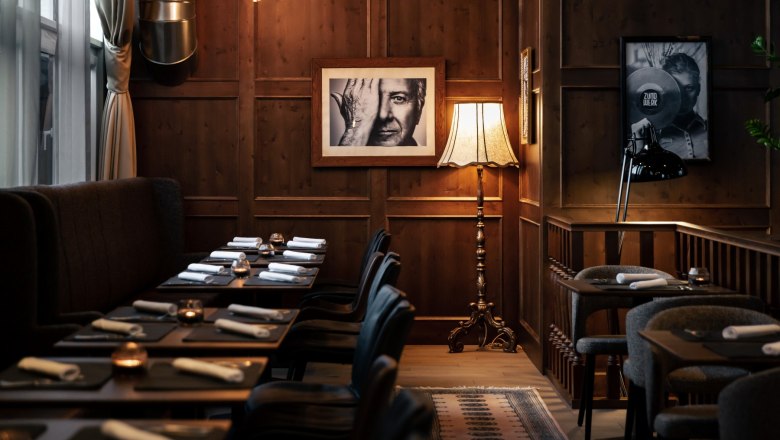 Living Room, © Derenko Design Gemütlicher Raum mit Holzwänden, gedeckten Tischen, Stehlampe und Schwarz-Weiß-Fotografien an der Wand.