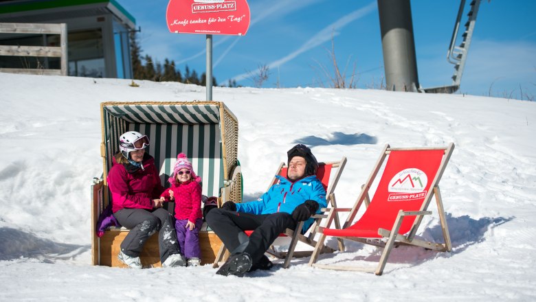 Genussplatz auf der Erlebnisalm, © Wiener Alpen/Ziegler Familie entspannt im Schnee auf der Erlebnisalm, sitzend in einem Strandkorb und Liegestühlen unter einem Schild mit der Aufschrift 'Genussplatz'.