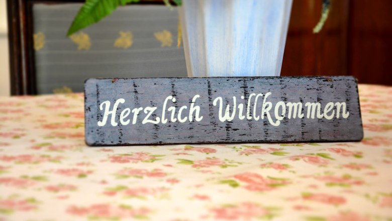 Herzlich Willkommen, © Gasthaus und Gästehaus Bsteh Ein Holzschild mit der Aufschrift 'Herzlich Willkommen' steht auf einem blumigen Tisch.