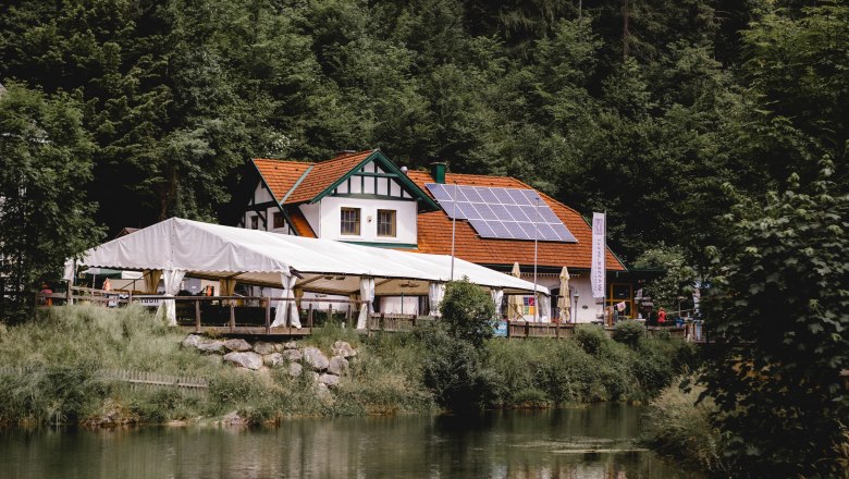 Gasthaus der Wander- & Wasserwelt Myrafälle, © Niederösterreich Werbung/David Schreiber Gasthaus mit Solarpanelen und Zelt am Waldrand, nahe einem Teich.
