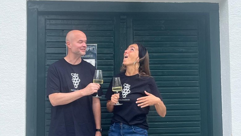 Heuriger "Die Neusiedlerin" Klein-Neusiedl, © Die Neusiedlerin Zwei Personen stoßen mit Weingläsern vor einem Gebäude mit dem Schild 'Neusiedlerin' an.