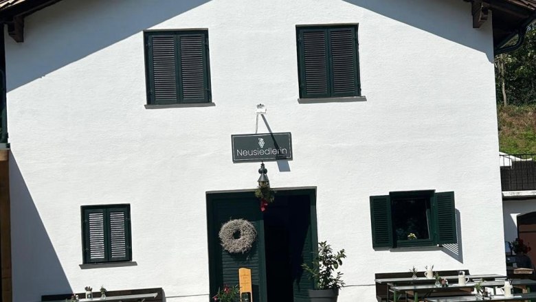 Heuriger "Die Neusiedlerin", Klein-Neusiedl, © Die Neusiedlerin Weißes Gebäude mit der Aufschrift 'Neusiedlerin' und Heurigentische davor.