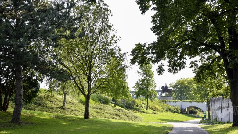 Grünräume in der Stadt, © Schwarzenegger Ein grüner Stadtpark mit Bäumen, einem Weg und einer Reiterstatue im Hintergrund.