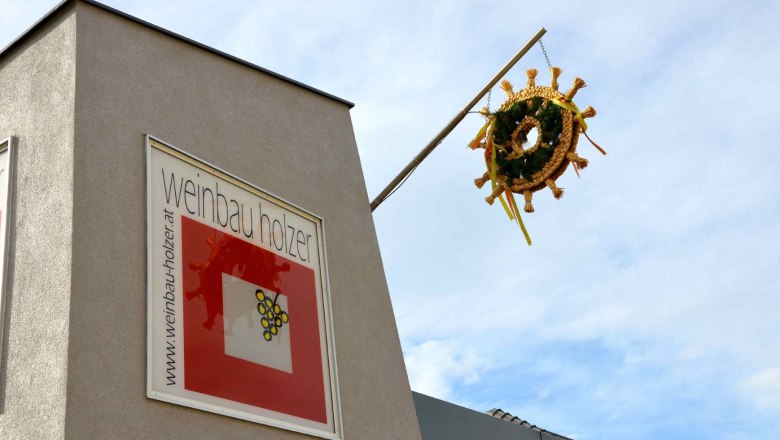 Weinbau Holzer, © Weinbau Holzer Gebäude mit Schild 'Weinbau Holzer' und einem dekorativen Kranz an einer Stange.