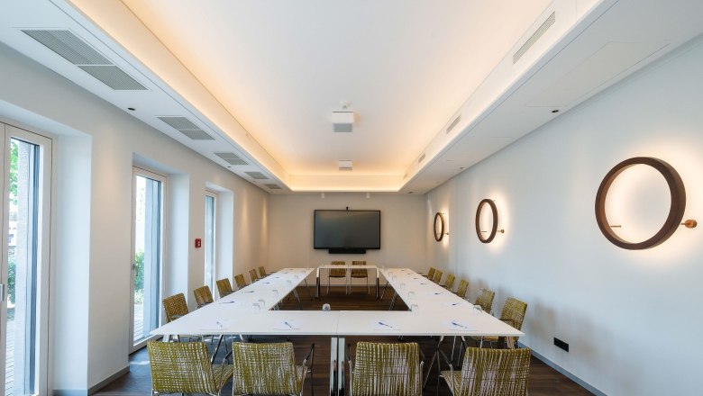 Der Seminarraum, © Philipp Blickfang Moderner Seminarraum mit U-förmiger Tischanordnung, Stühlen, einem großen Bildschirm und dekorativen Wandlampen.