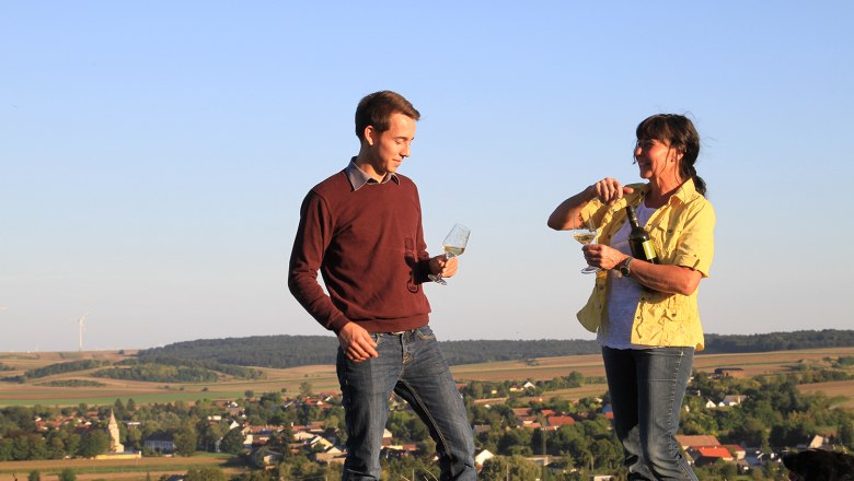 Weingut Thüringer, © Weingut Thüringer Zwei Personen stehen auf einem Hügel mit Weingläsern in der Hand, im Hintergrund eine ländliche Landschaft mit Windrädern.
