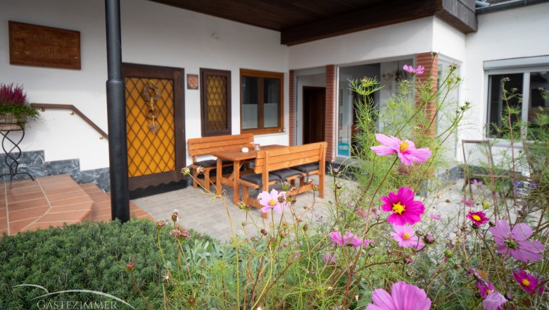 Terrasse, © Doris Eminger Terrasse mit Holzmöbeln und blühenden Blumen im Vordergrund.