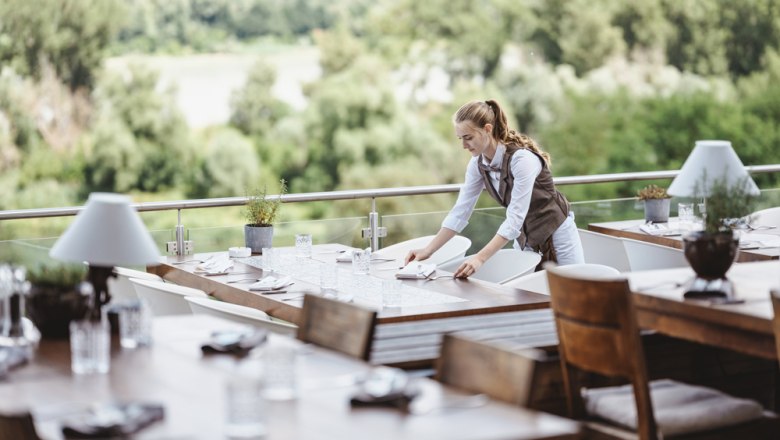 Ausblick auf den Nationalpark Donau-Auen, © Niederösterreich Werbung/Michael Reidinger Eine Kellnerin deckt Tische auf einer Terrasse mit Blick auf den Nationalpark Donau-Auen.