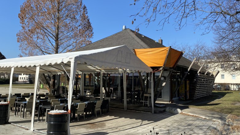 Mitten im Eduard-Garderer-Park, © Wiener Alpen Ein Café mit Außensitzbereich im Eduard-Garderer-Park, umgeben von kahlen Bäumen und blauem Himmel.