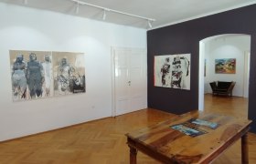 die Galerie MERKIMA, © Doris Hauser Innenansicht einer Kunstgalerie mit Holzboden, Gemälden an den Wänden und einem Holztisch in der Mitte.