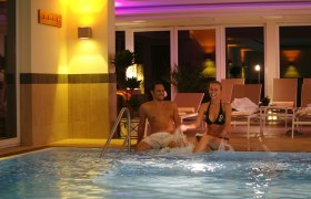 Indoor-Pool im Wienerwaldhof, © Wienerwaldhof Ein Mann und eine Frau sitzen am Rand eines beleuchteten Indoor-Pools und lachen. Im Hintergrund sind Liegestühle und Pflanzen zu sehen.