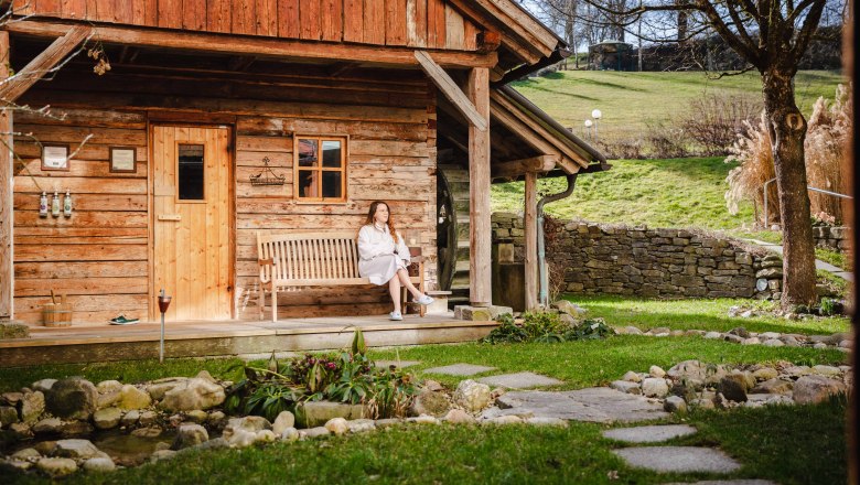 RelaxResort Kothmühle, © Johanna Meinschad Frau in Bademantel sitzt auf einer Bank vor einer Holzhütte im Grünen.