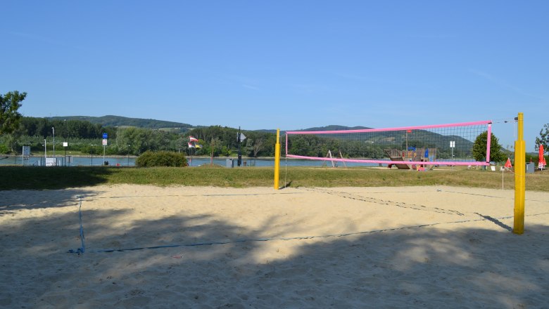 Beachvolleyplatz, © Stadtgemeinde Korneuburg Ein leerer Beachvolleyballplatz mit gelben Pfosten und rosa Netz vor einem Fluss und grünen Hügeln im Hintergrund.