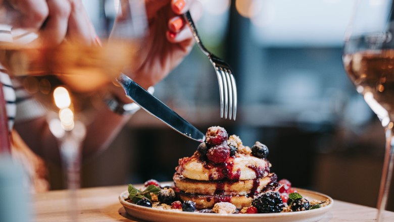 Speisenbeispiel Pier 4, © Pier 4 Nahaufnahme von Pancakes mit Beeren und Sirup, eine Person schneidet mit Messer und Gabel.