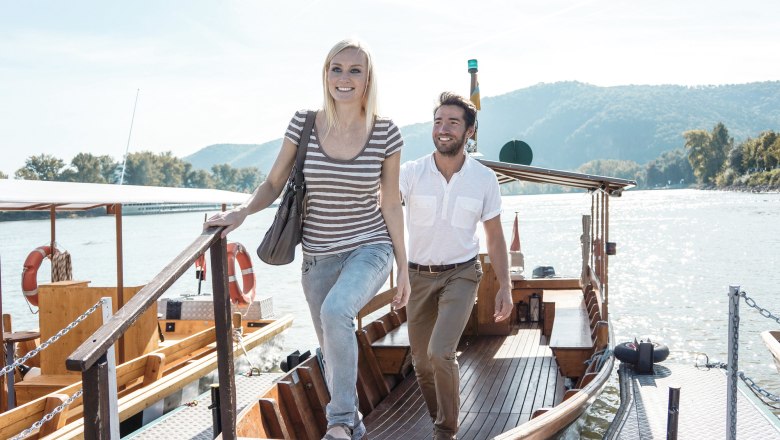 Autofrei unterwegs in der Wachau, © Donau Niederösterreich/Steve Haider Ein Mann und eine Frau steigen von einem Boot auf einen Steg. Im Hintergrund ist ein Fluss und bewaldete Hügel zu sehen.