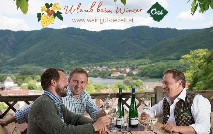 Winzer Gespräche, © RN Drei Männer sitzen an einem Tisch mit Weinflaschen und Gläsern, im Hintergrund eine malerische Landschaft mit Hügeln.