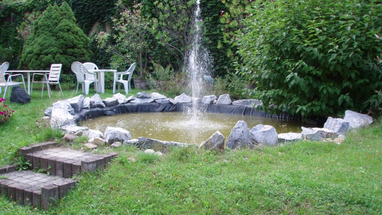 Hotel Wegscheidhof, © Hotel Wegscheidhof Garten mit kleinem Teich und Springbrunnen, umgeben von Pflanzen und Gartenmöbeln.