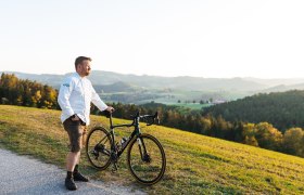 Wirt Andreas Ottner ist passionierter Biker, © Niederösterreich Werbung/Mara Hohla Ein Mann in weißem Hemd steht mit einem Fahrrad auf einem Hügel mit Blick auf eine grüne Landschaft.