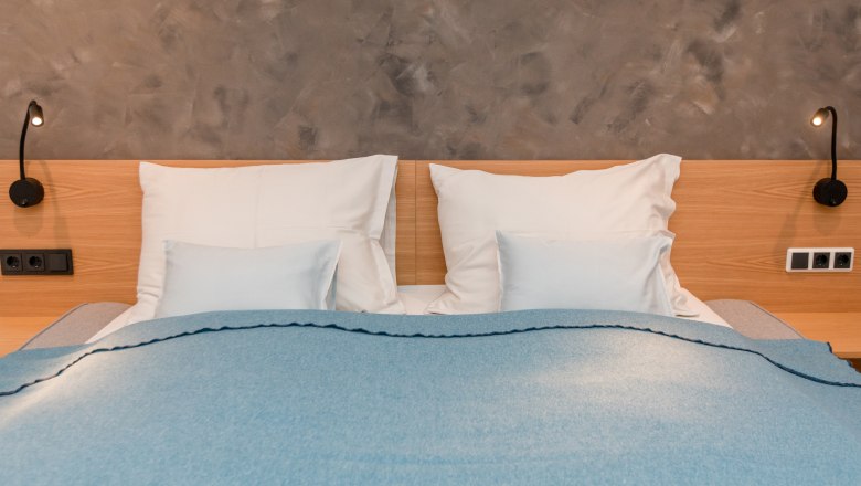 Haus am See, © schwarz-koenig.at Ein Doppelbett mit blauer Decke und weißen Kissen vor einer grauen Wand.