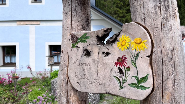 Kräutergarten Kanlehen, © "Natur im Garten" Holzschild mit Blumenmalerei und Schriftzug 'Willkommen im Kräutergarten' vor einem blauen Gebäude.
