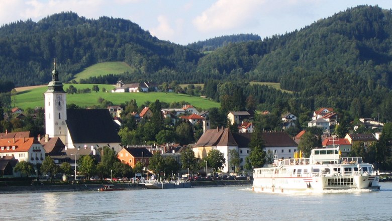 Anlegestelle Grein 3 in Grein, © Donau Schiffsstationen GmbH Stadt Grein mit Kirche und Schiff auf der Donau, umgeben von Hügeln.
