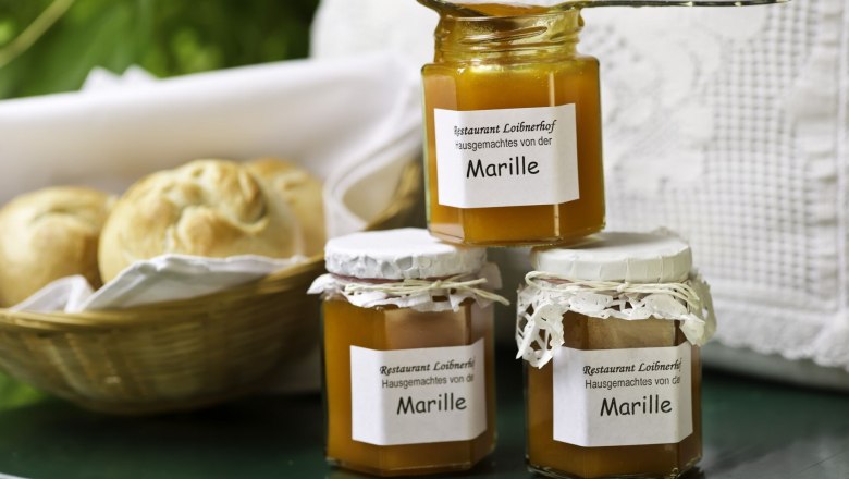 Produkte vom Loibnerhof, © Rita Newman Drei Gläser Marillenmarmelade vom Restaurant Loibnerhof neben einem Korb mit Brötchen.