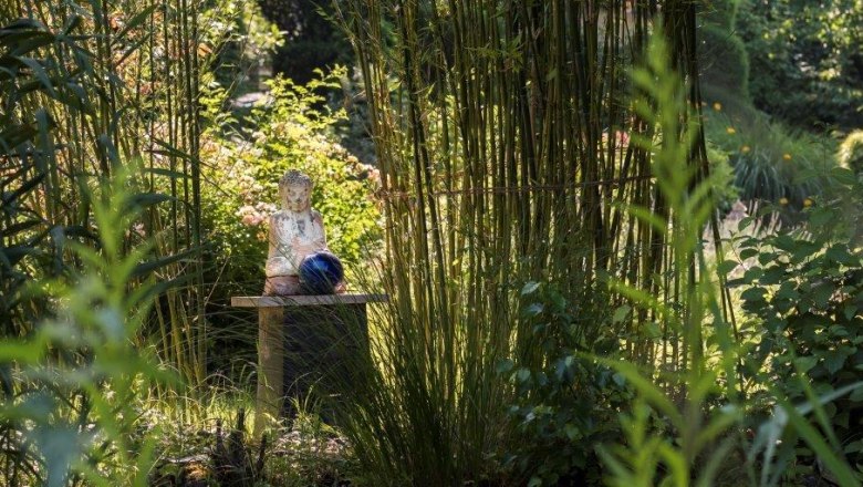 Asienflair in Rosenbauchs Restaurantgarten, © Karl Rosenbauch Ein asiatisch inspirierter Garten mit einer Buddha-Statue auf einem Tisch, umgeben von Bambus und üppigem Grün.