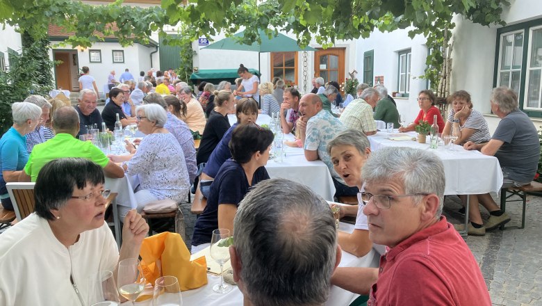 Unterhaltsame Stimmung im Winzerhof der Familie Ruttenstock, © Weingut Ruttenstock Menschen sitzen an Tischen im Freien unter Weinreben und unterhalten sich.