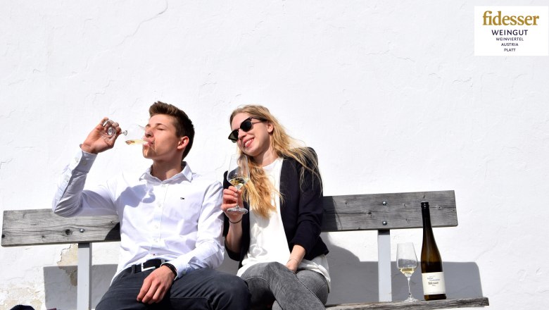 Sophie und Rudolf Fidesser, © Weingut Fidesser Zwei Personen sitzen auf einer Bank und trinken Wein vor einer weißen Wand.