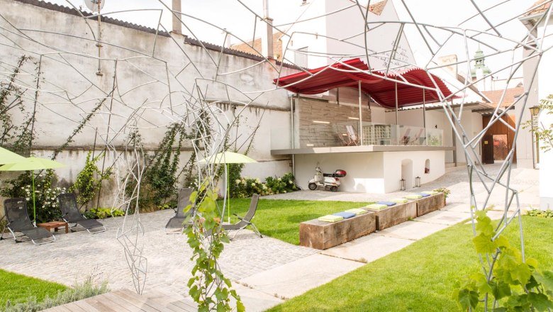 Klinger's Gästehaus, © Astrid Bartl Moderner Innenhof mit Rasen, Liegestühlen, Metallskulpturen und einer Vespa unter einem roten Vordach.