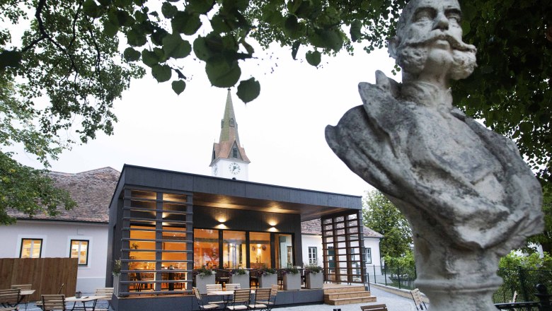 Das FRANZ JOSEPH Wirtshaus am Kaiserplatz, © Astrid Bartl Büste vor modernem Gebäude mit Kirchturm im Hintergrund, umgeben von Bäumen.