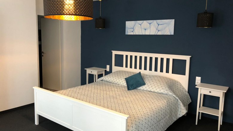 Doppelzimmer, © Piestingerhof Ein modernes Doppelzimmer mit einem weißen Bett, blauer Wand und stilvollen Lampen.